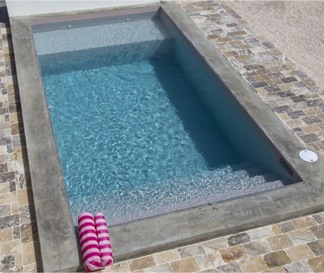 Petite piscine rectangulaire remplie d’eau claire, avec revêtement Aquabright gris, marches immergées et serviettes roses sur la bordure.