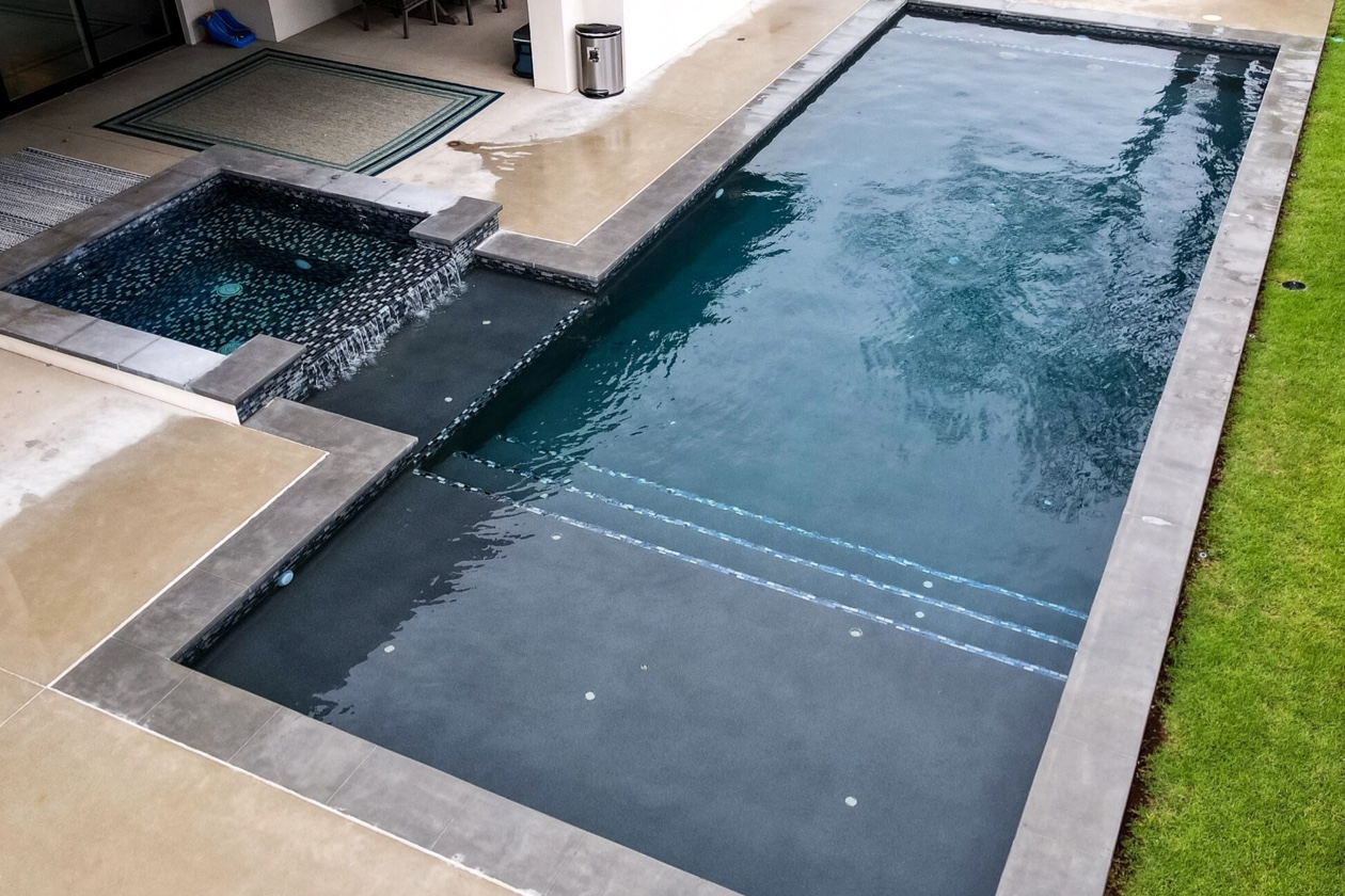 Piscine résidentielle moderne au fini Aquabright gris foncé, avec large escalier immergé, spa intégré et cascade sur le côté.