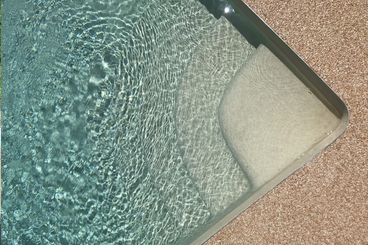 Vue rapprochée d’une marche arrondie dans une piscine au revêtement Aquabright gris clair, sous une eau limpide bordée d’une terrasse en granulat.