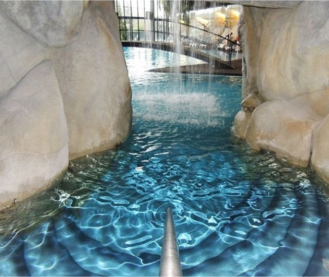 Passage aquatique intérieur entre des parois rocheuses, avec cascade, rampe centrale, marches immergées et revêtement Aquabright bleu profond.