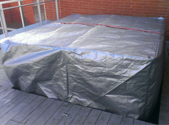 Outdoor spa covered with a grey protective cover held in place by a red cord, on a wooden deck after being closed for winter.