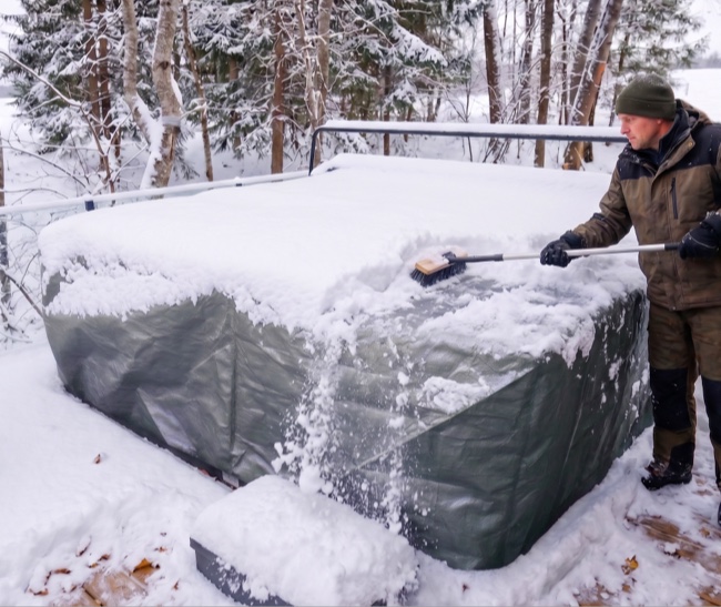 Man using a broom to clear snow accumulated on the protective cover of a spa closed for winter, in the middle of a snow-covered yard.